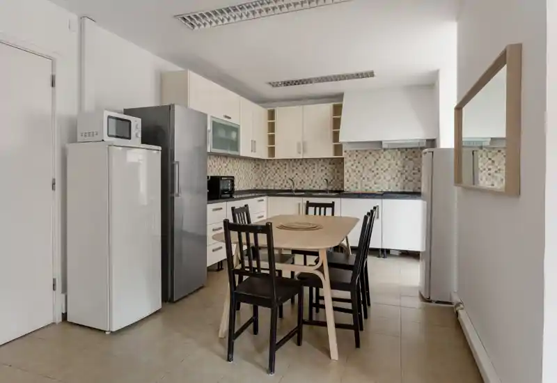 Bright modern kitchen with a central wooden dining table and four chairs. Light cabinetry, mosaic backsplash and full-size refrigerator create a functional, tidy cooking and eating space.