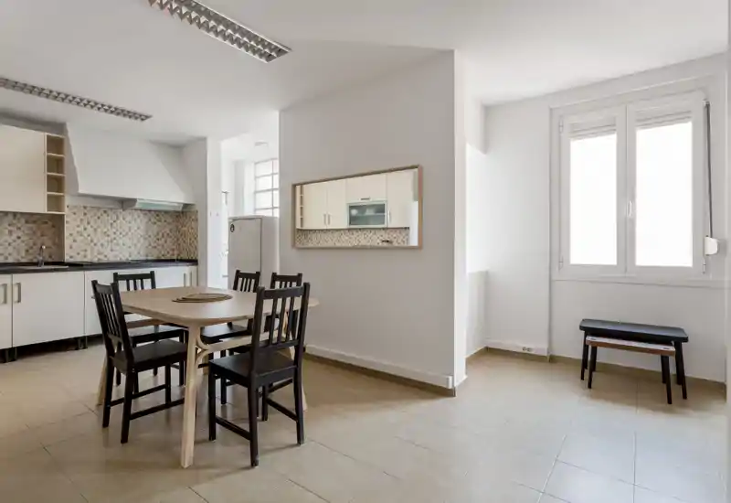 Bright, minimalist dining area integrated with a compact kitchen. A wooden table with four chairs sits centrally, with neutral tiles and a large window providing natural light.