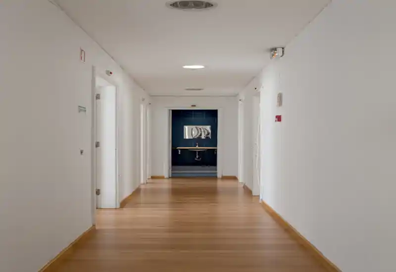 A clean, minimal interior corridor with wooden flooring leading to a sink area at the far end. White walls, recessed ceiling lights and several doorways create a neutral, well-lit passage ideal for apartment circulation.