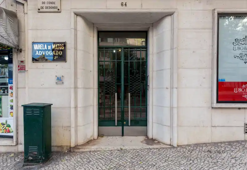 Street-level building entrance with a green metal double door set in light stone cladding; mosaic pavement in front and signage beside the doorway.