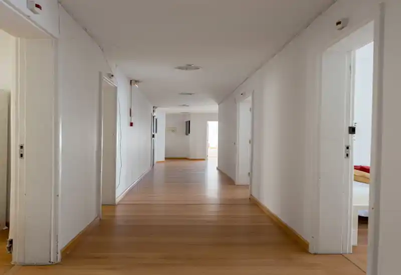 Long, bright interior corridor with wooden flooring and multiple doorways leading to adjacent rooms; neutral white walls and ceiling lights create a clean, simple entrance/hallway space.