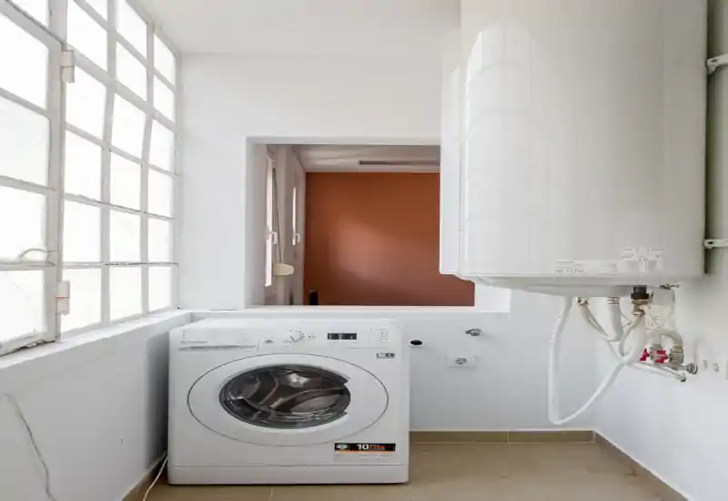 Small, bright utility/laundry room with a front-load washing machine and a wall-mounted water heater. The space is clean, well-lit by a large glass-block window and has a neutral tile floor.