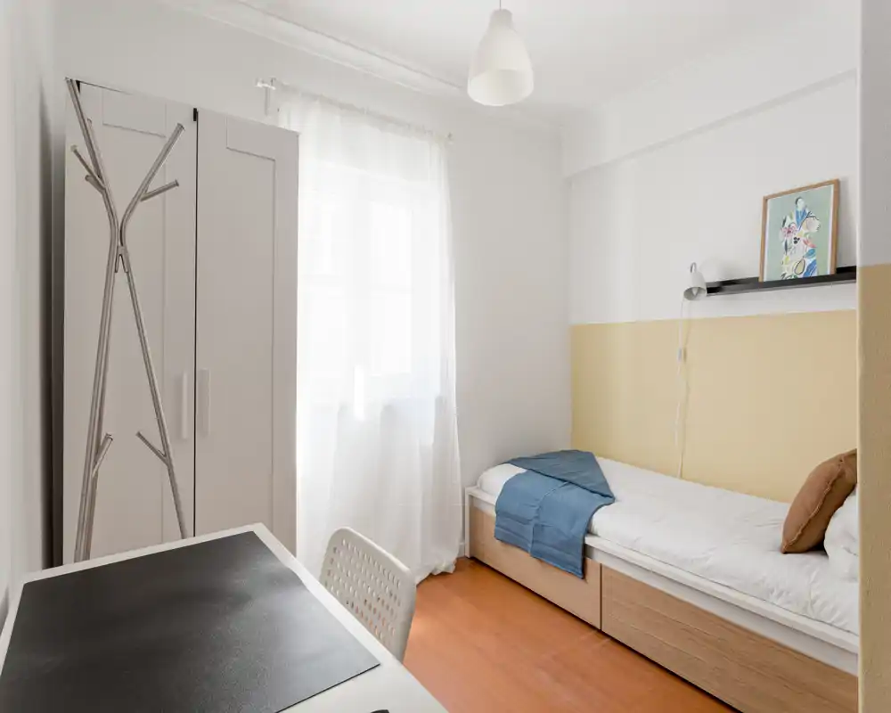 Chambre simple lumineuse et bien mise en scène avec lit bas, tiroirs de rangement sous le lit et fenêtre laissant entrer une lumière douce. Décor minimal de style scandinave avec armoire, bureau et textiles chaleureux.