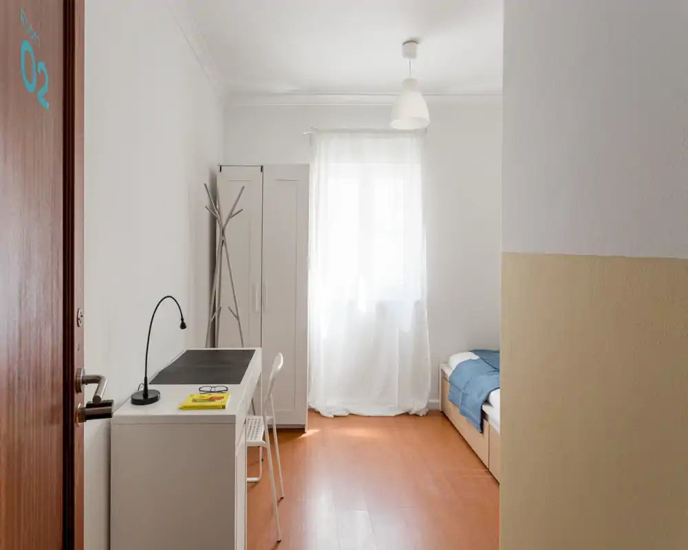 Chambre simple lumineuse et minimaliste avec lit bien fait, bureau et armoire près d'une fenêtre laissant entrer une douce lumière naturelle — idéale pour étudiant ou voyageur solo.