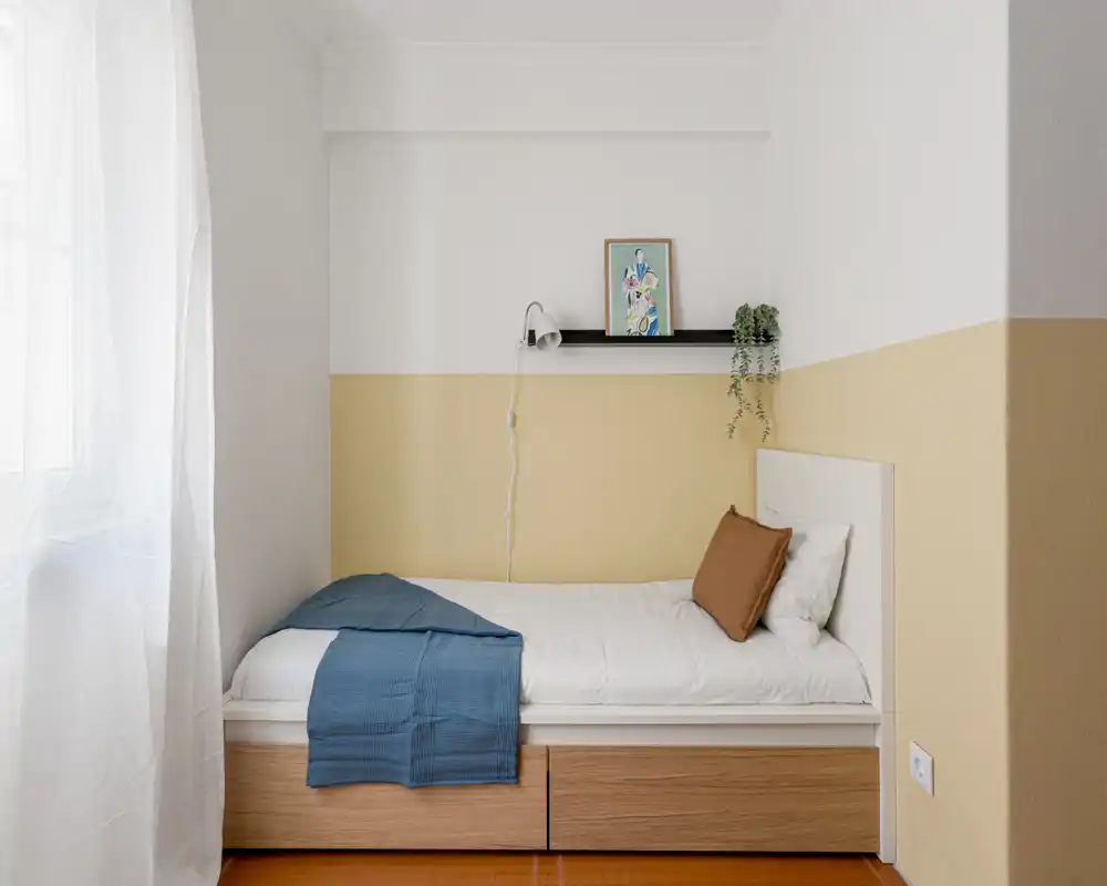 Petite chambre lumineuse avec lit simple, rangement en bois sous le lit, linge de lit sobre et une étagère décorative au-dessus. La lumière naturelle de la fenêtre apporte une ambiance calme et accueillante, idéale pour une personne.