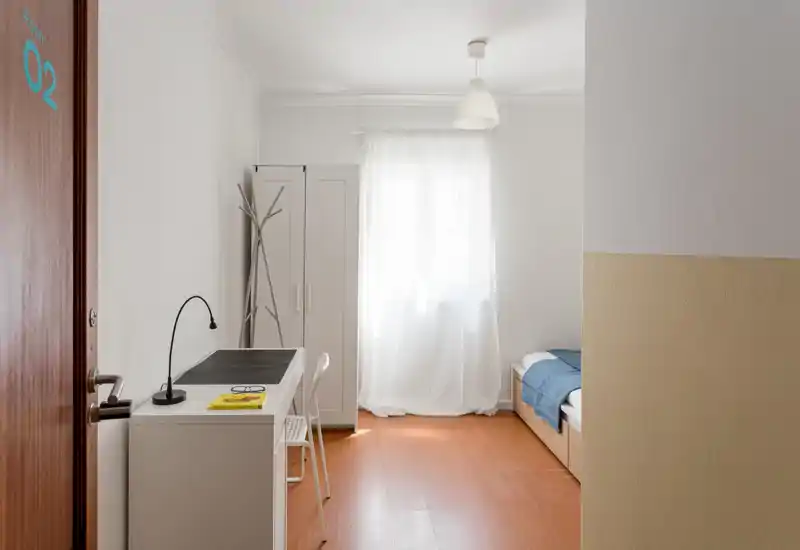 Chambre simple lumineuse et minimaliste avec lit bien fait, bureau et armoire près d'une fenêtre laissant entrer une douce lumière naturelle — idéale pour étudiant ou voyageur solo.