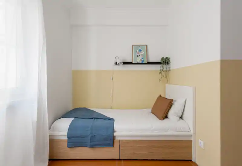 Petite chambre lumineuse avec lit simple, rangement en bois sous le lit, linge de lit sobre et une étagère décorative au-dessus. La lumière naturelle de la fenêtre apporte une ambiance calme et accueillante, idéale pour une personne.