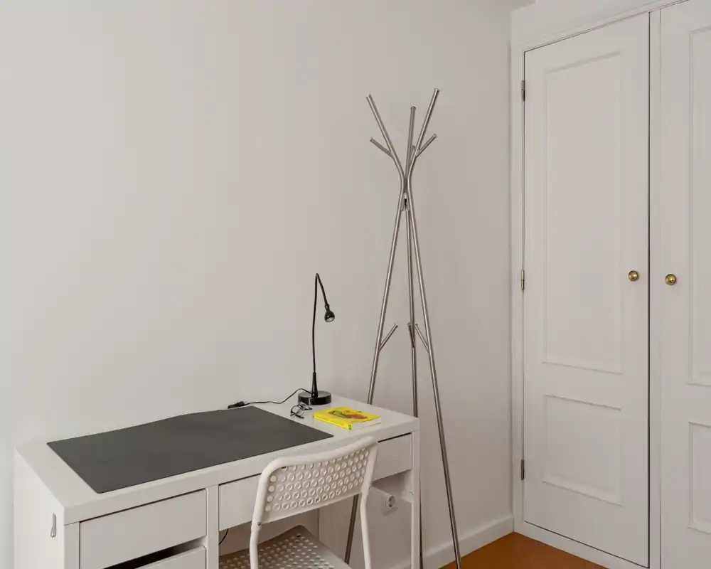 Coin minimaliste et lumineux avec bureau blanc, chaise perforée et porte-manteau fin ; espace de travail soigné idéal pour le télétravail ou l'étude.