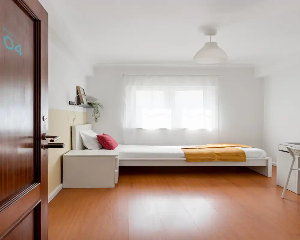 Chambre simple lumineuse et minimaliste avec lit bas blanc, table de chevet et bureau — idéale pour une location épurée et aérée. La lumière naturelle et le sol en bois apportent une ambiance chaleureuse.