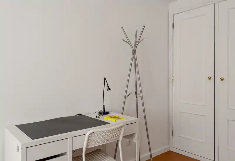 Coin minimaliste et lumineux avec bureau blanc, chaise perforée et porte-manteau fin ; espace de travail soigné idéal pour le télétravail ou l'étude.