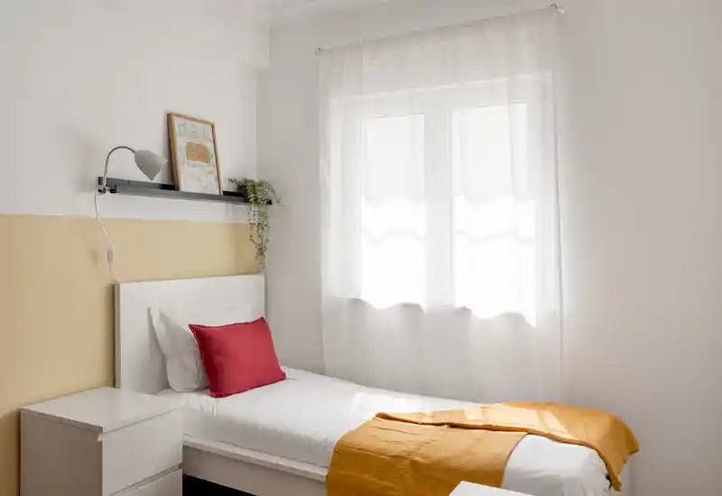 Chambre simple lumineuse et soignée avec lit blanc, plaid et coussin colorés, éclairée naturellement par une fenêtre aux voilages légers.