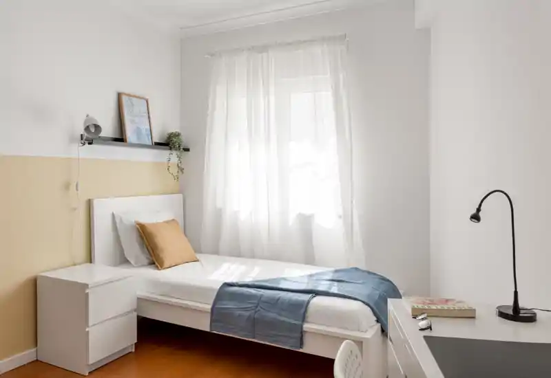 Chambre simple lumineuse et minimaliste avec un lit bien fait, tissus clairs et un petit bureau. Grande fenêtre avec voilages laissant entrer une belle lumière naturelle.