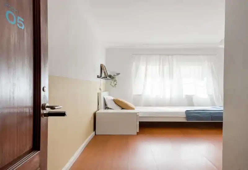 Chambre simple lumineuse et minimaliste avec lumière naturelle, lit bas blanc et table de chevet, sol en bois chaleureux et textiles doux pour une ambiance calme.
