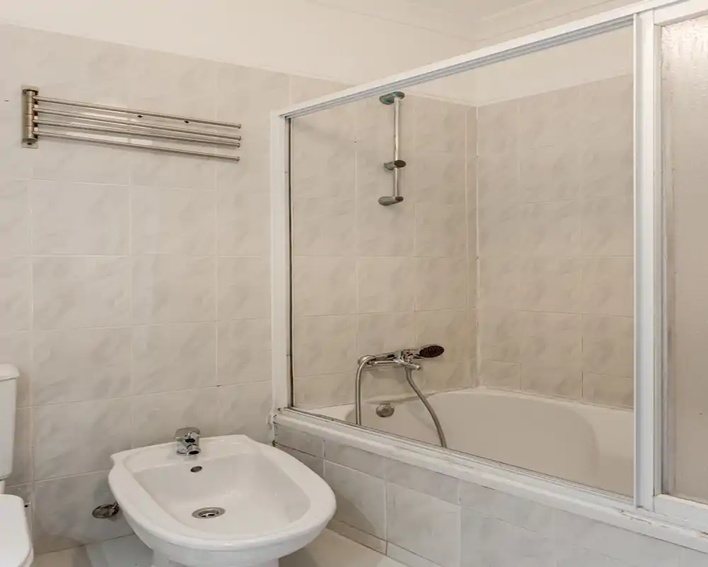 Salle de bain carrelée propre avec baignoire et douche, bidet et WC visibles. Carrelage neutre et paroi vitrée pour un espace pratique et bien éclairé.