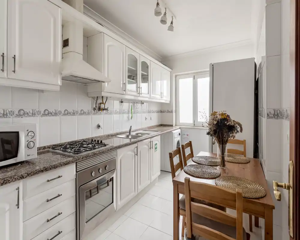 Cuisine claire et bien équipée avec placards blancs, plan de travail en granit, four encastré et une petite table en bois — idéale pour des repas conviviaux.