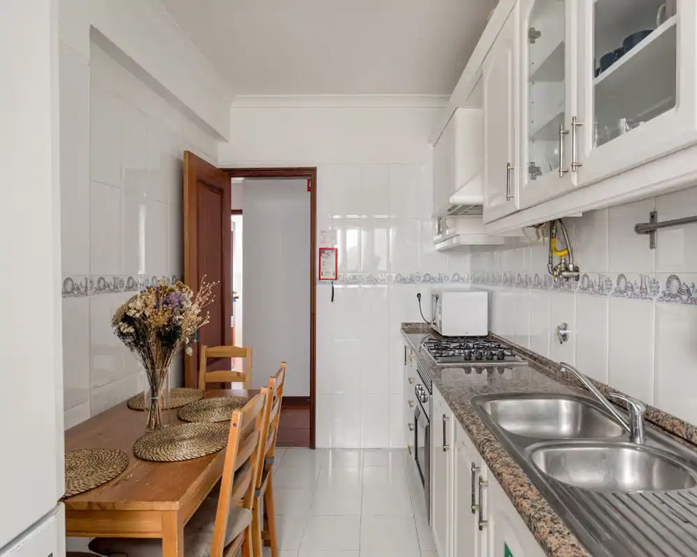Cuisine étroite et lumineuse avec meubles blancs, plan de travail en granit, double évier et petite table à manger avec chaises — idéale pour les petits logements.