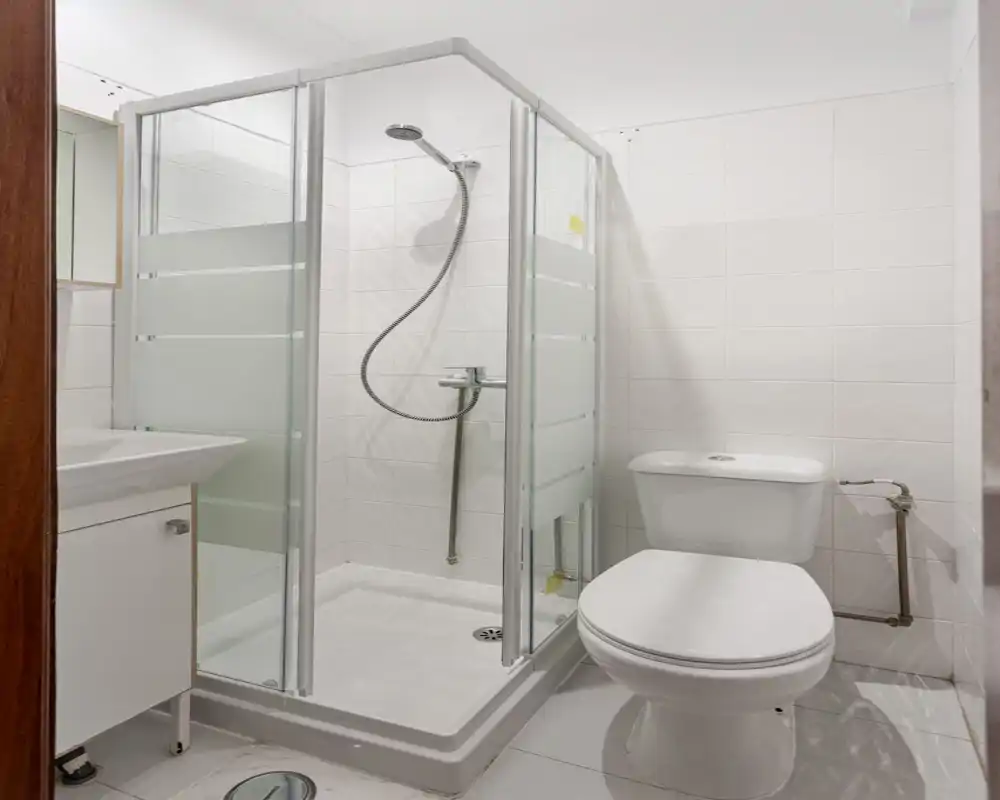 Salle de bains propre et compacte avec cabine de douche d'angle, WC et lavabo ; carreaux blancs lumineux et équipements modernes pour un espace soigné et fonctionnel.