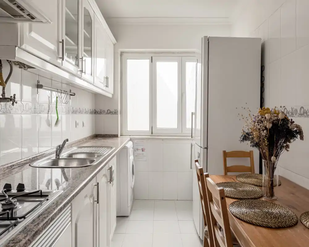 Cuisine lumineuse et propre de type couloir avec plans de travail effet marbre, plaque à gaz intégrée, évier, réfrigérateur et petite table en bois avec sets de table décoratifs et fleurs séchées.