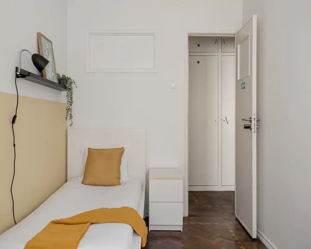 Tidy single bedroom with a white headboard, mustard cushion and throw, a nightstand and clean parquet flooring — bright, minimal and welcoming.