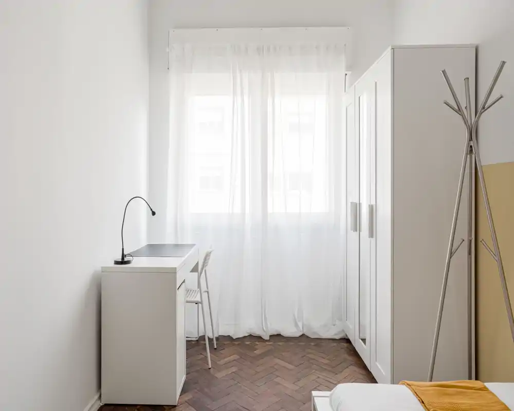 Minimal, bright bedroom corner with a small desk, wardrobe and a partially visible bed with warm throw. Clean, airy styling ideal for a single occupant.