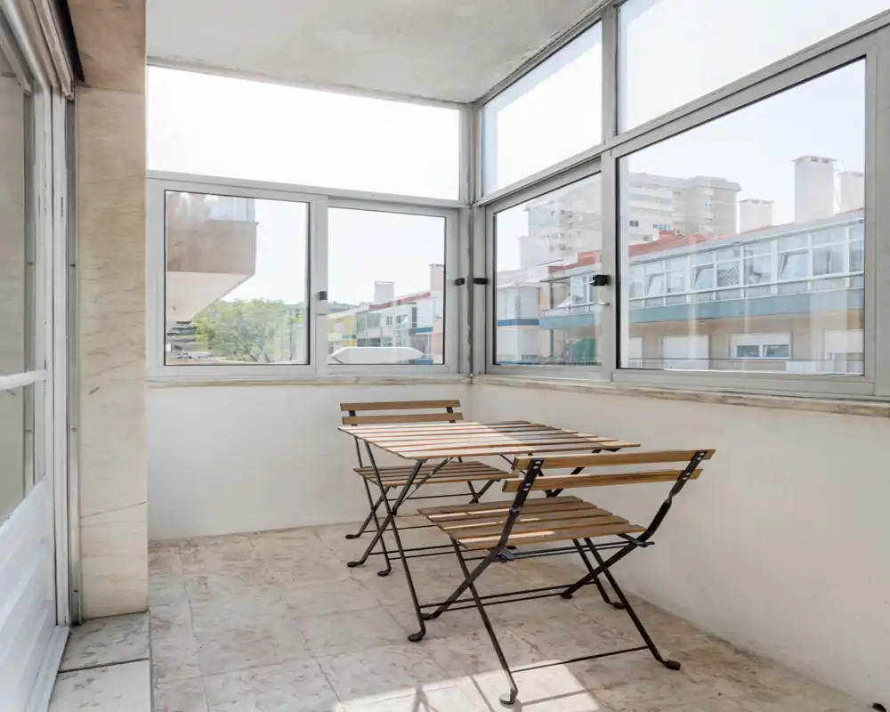 Bright enclosed balcony with large windows and a simple wooden folding table and chairs — ideal for morning coffee or a small outdoor seating area.