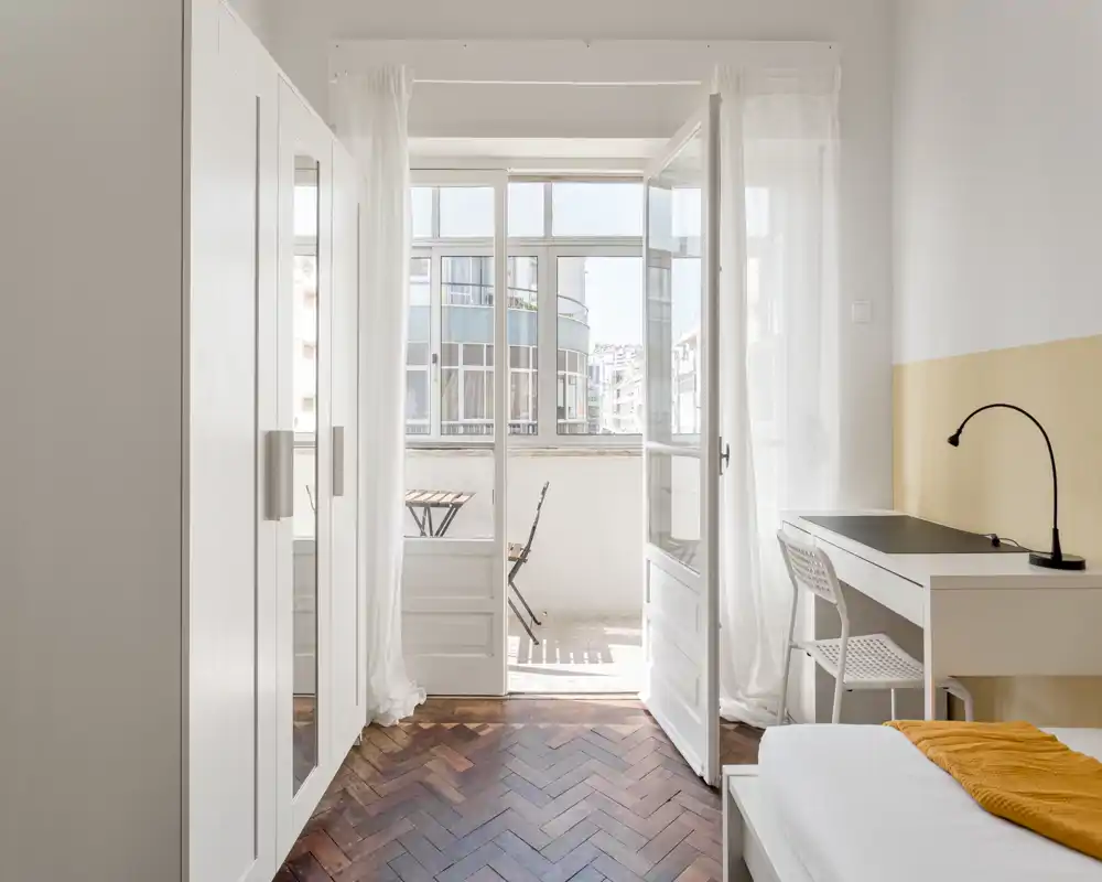 Bright, minimal bedroom with parquet flooring, a small desk and a balcony accessible through glass doors — a cozy, well-lit space ideal for single occupancy.