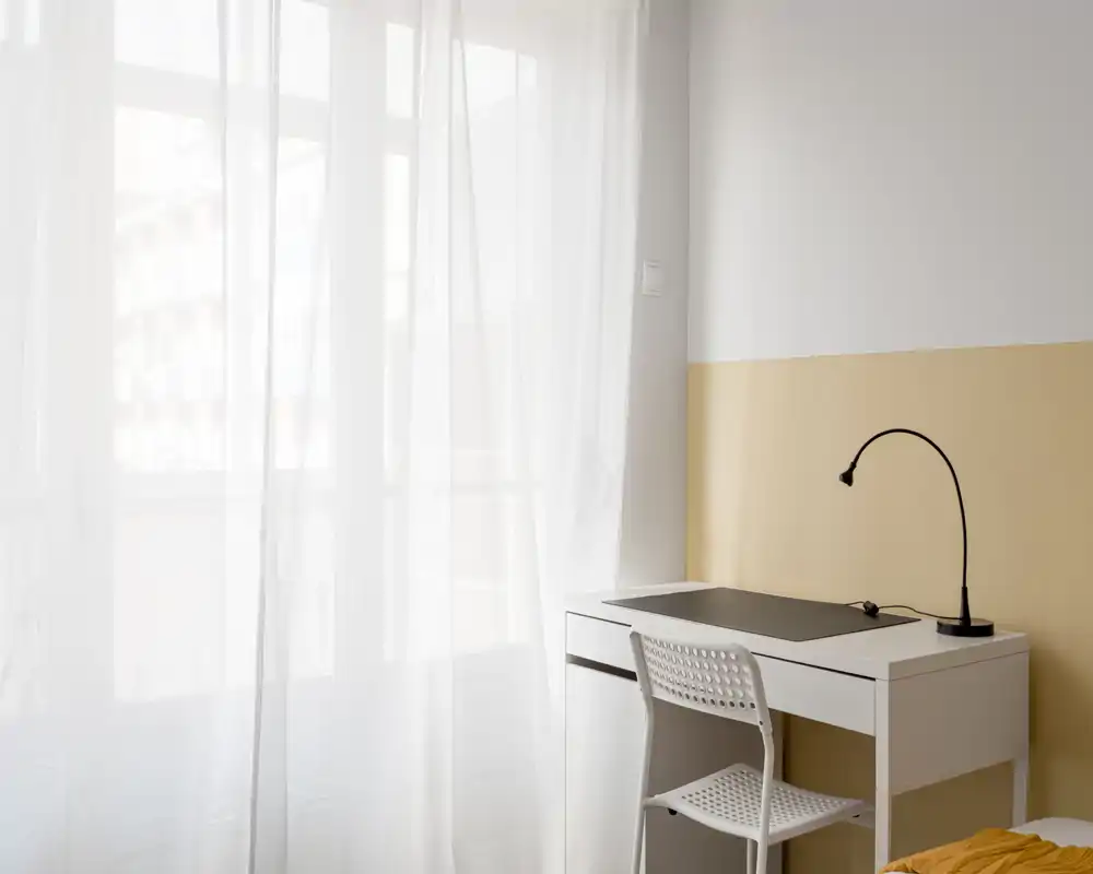 Bright, minimal bedroom corner with a white desk, perforated chair and a flexible black desk lamp beside sheer curtains that diffuse natural light.