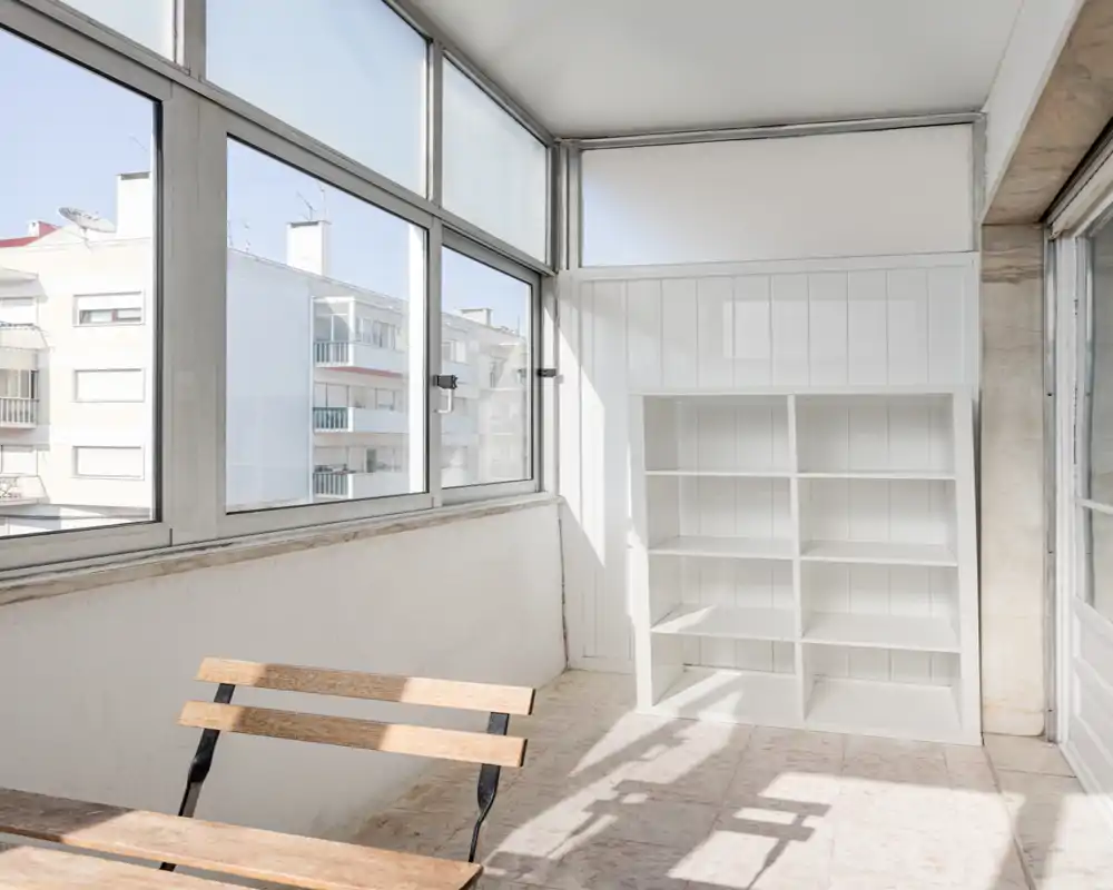 Bright enclosed balcony / sunroom with large windows, a simple wooden bench and built-in shelving — ideal for a small reading nook or storage area flooded with natural light.
