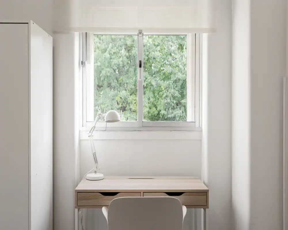 Bright, minimal workspace by a window: a clean wooden desk with a lamp and chair positioned under a large leafy window, ideal for focused work.