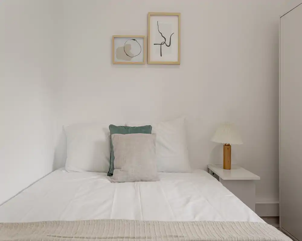 Bright, minimalist bedroom with a neatly made double bed, soft neutral textiles and simple wall art — ideal for a calm rental listing.