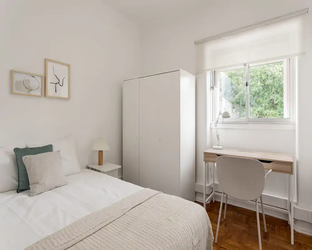 Bright, minimalist bedroom with a comfortable double bed, bedside table and a wardrobe next to a windowed workspace — fresh neutral tones and natural light.