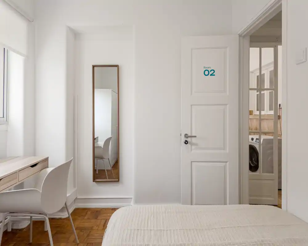 Bright, minimal bedroom with a visible bed, wooden parquet floor, a desk by the window and a tall mirror — clean, airy and well-lit.