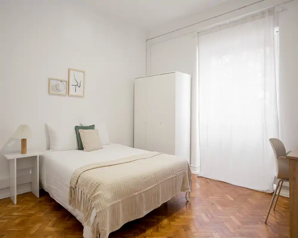 Bright, minimalist bedroom with a neatly made double bed, soft neutral linens, a wardrobe and a desk by a large window that floods the room with natural light.