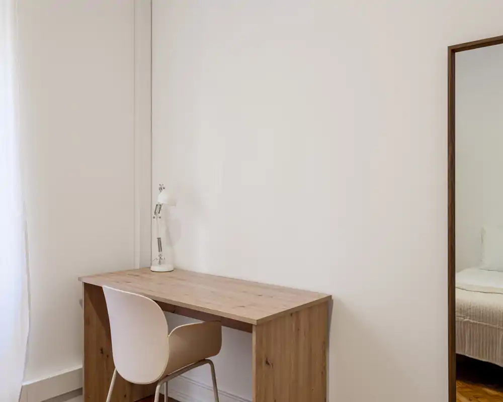 Small bedroom corner shown with a wooden desk and chair; the bed is partially visible in a mirror at the right edge. Bright, minimalist styling with neutral tones and natural light.