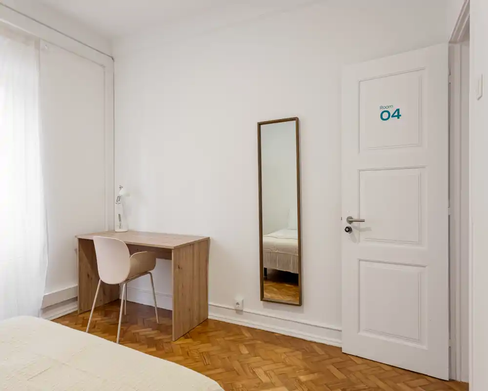 Bright, minimal bedroom with a visible bed corner, wooden parquet floor, a simple desk and a full-length mirror — ideal for a clean, modern rental listing.