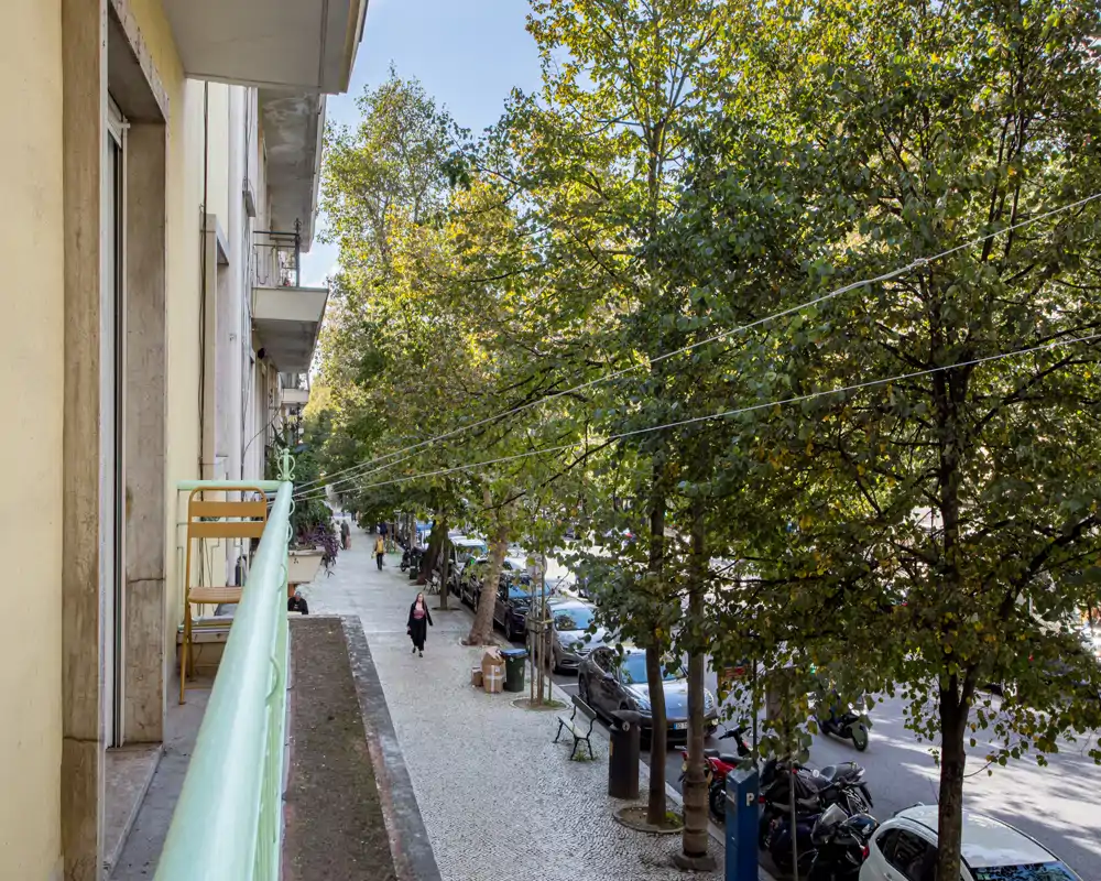 Sunny balcony view overlooking a tree-lined city sidewalk and parked cars, with a small railing and outdoor chairs — ideal to showcase an apartment’s outdoor space and street atmosphere.