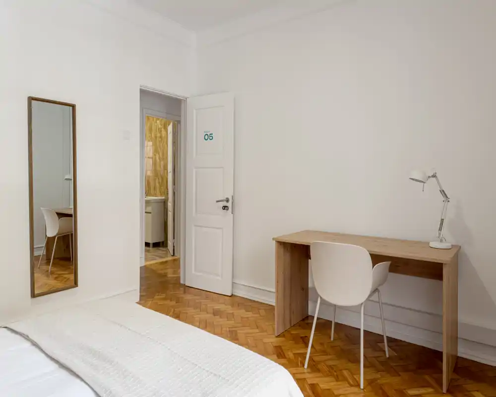 Minimal, bright bedroom with a visible bed corner, wooden parquet floor, a simple desk and chair, and a full-length mirror by the wall — ideal for a calm, uncluttered stay.