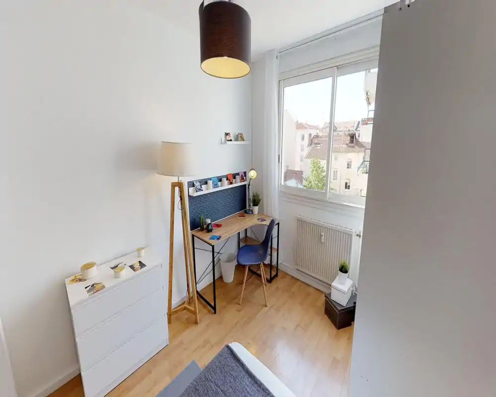 Coin de chambre lumineux et bien mis en scène avec le bord du lit visible, sol en bois, bureau près de la fenêtre et lampes et plantes qui apportent une ambiance chaleureuse.