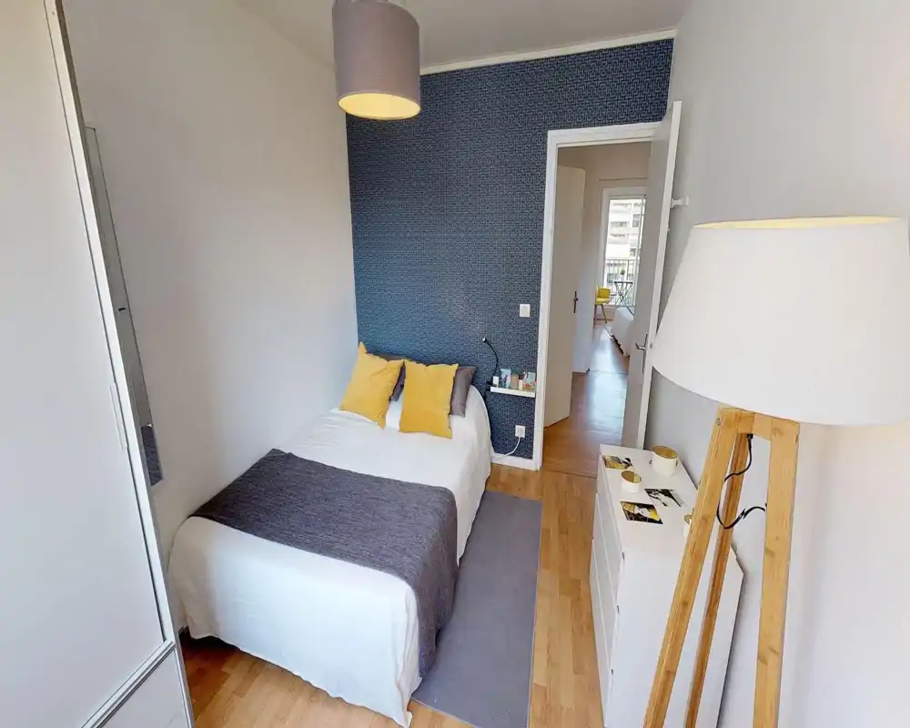 Chambre lumineuse et soignée avec un lit double en linge blanc, plaid gris et coussins jaunes. Un mur d'accent bleu, parquet et éclairage doux donnent une ambiance moderne et chaleureuse.