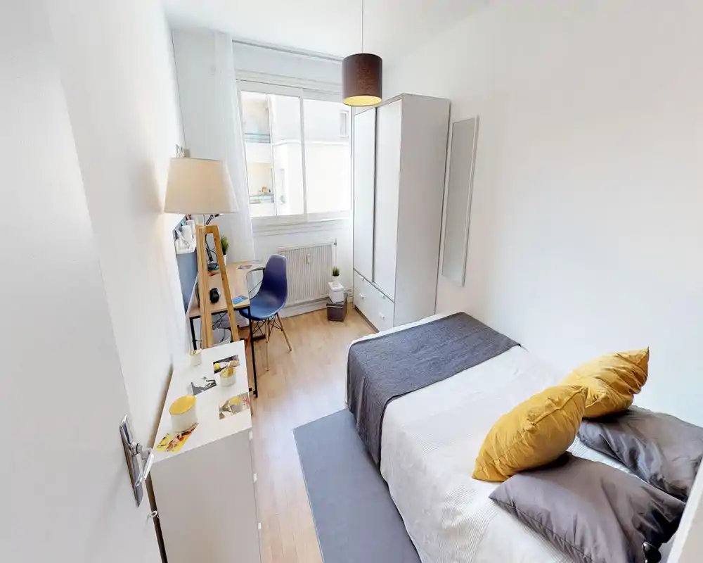 Chambre simple lumineuse et bien aménagée avec lit fait, coussins jaunes, petit bureau près de la fenêtre et armoire blanche — parfaite pour un étudiant ou un professionnel.