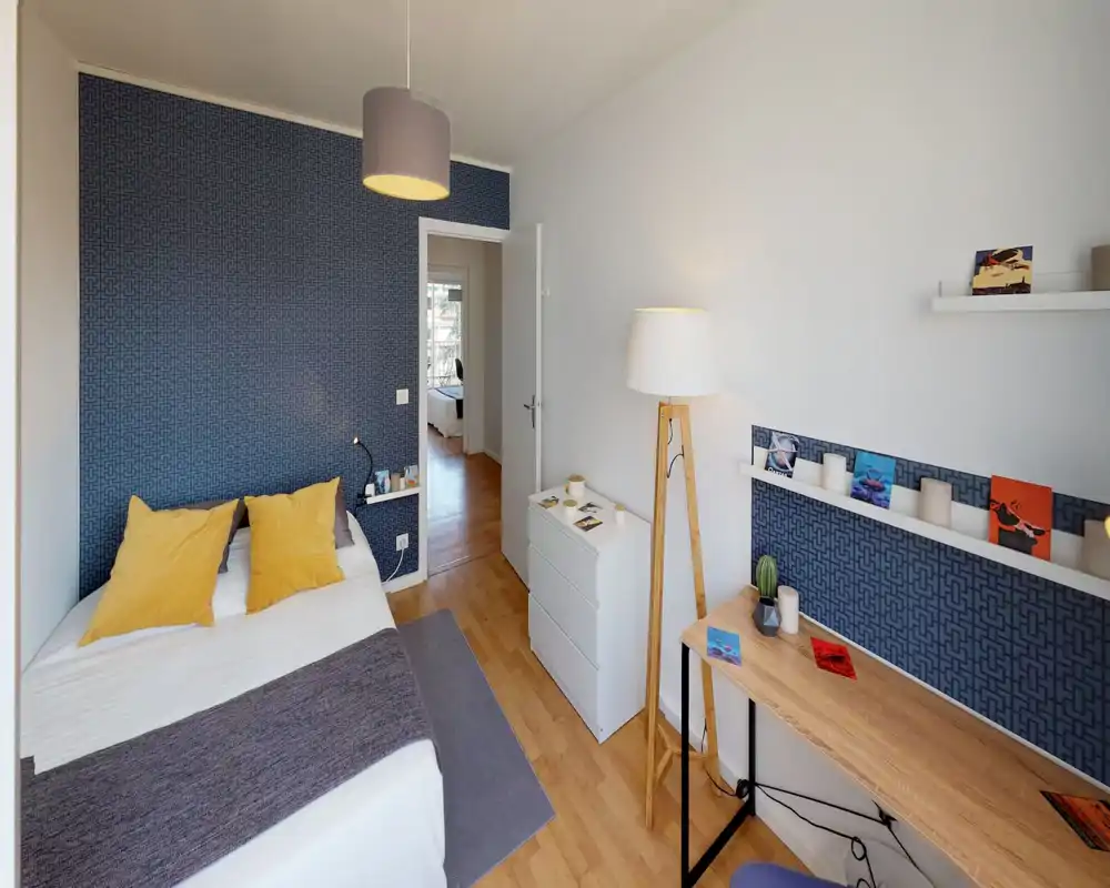 Chambre lumineuse et soignée avec un lit complet, coussins jaunes et un mur d'accent bleu à motifs; un bureau en bois et un éclairage doux apportent un coin travail chaleureux.
