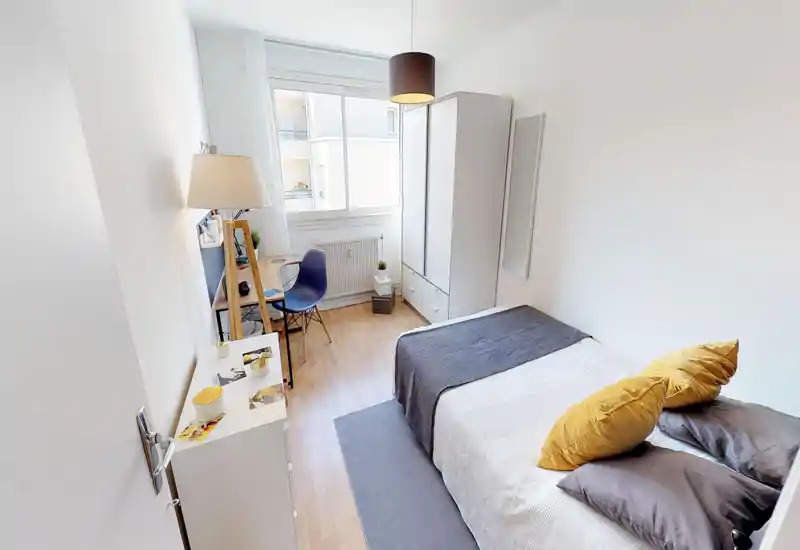 Camera da letto singola luminosa e ben arredata con letto ordinato, cuscini gialli, una scrivania vicino alla finestra e un armadio bianco — ideale per studente o professionista.