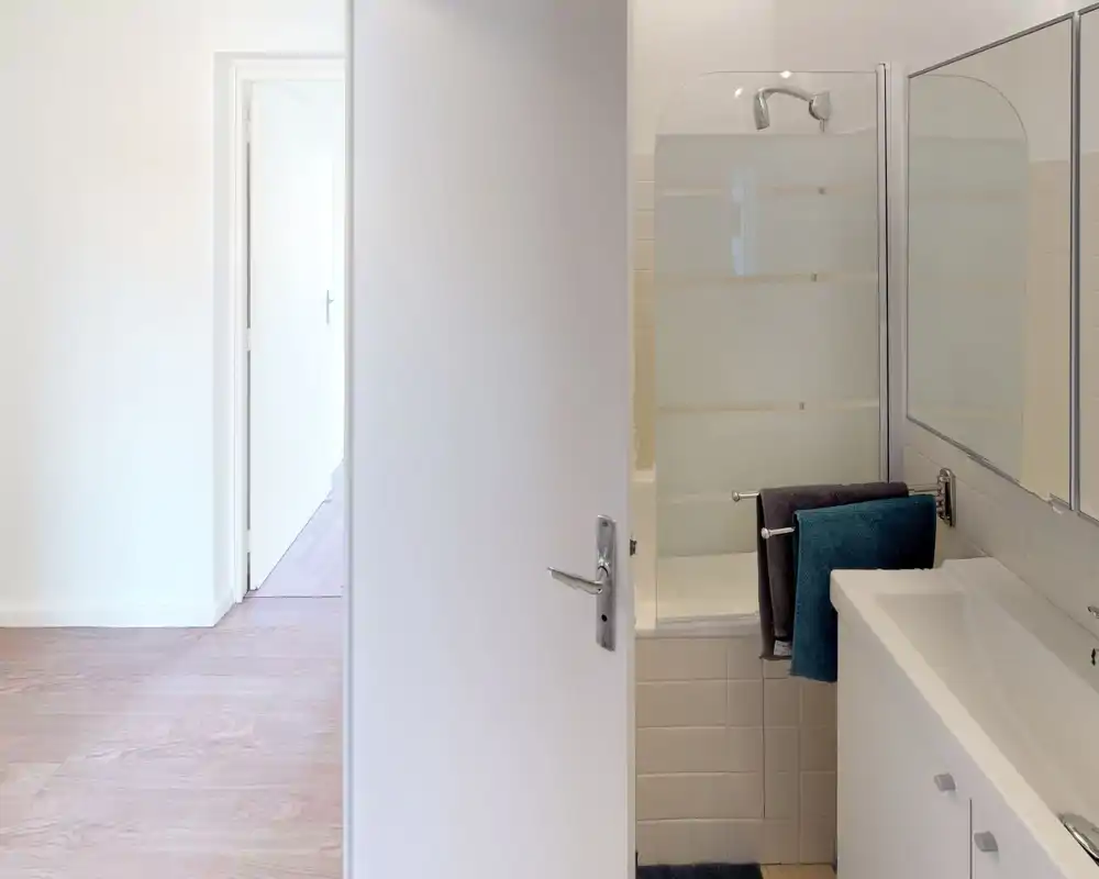 Salle de bains compacte avec lavabo, miroir et baignoire équipée d'un pare-douche en verre dépoli ; carrelage neutre et style propre et minimaliste.