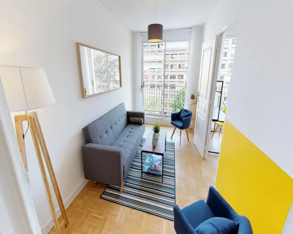 Salon moderne et lumineux avec canapé gris, tapis rayé et fauteuils bleus autour d'une petite table basse; grande fenêtre avec balcon apportant une belle lumière naturelle.