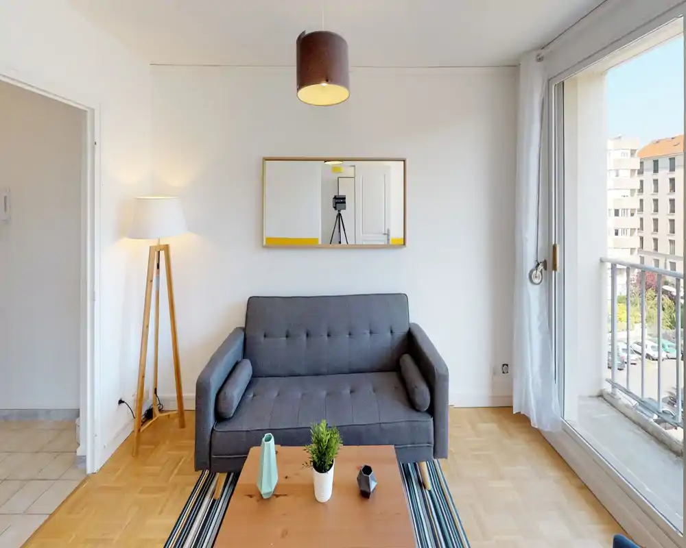 Salon moderne et lumineux avec canapé gris compact, lampadaire, table basse et grande baie vitrée ouvrant sur un balcon qui inonde la pièce de lumière naturelle.
