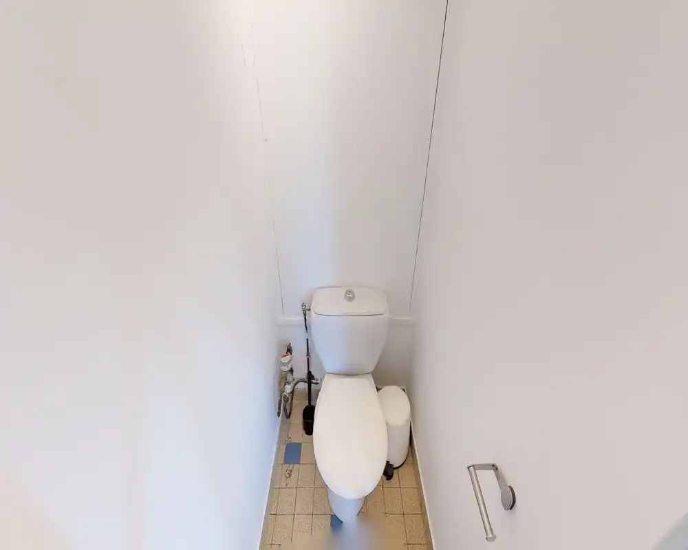 Toilettes compactes avec WC standard, sol carrelé et murs blancs ; espace simple et propre, adapté comme toilettes d'appoint.