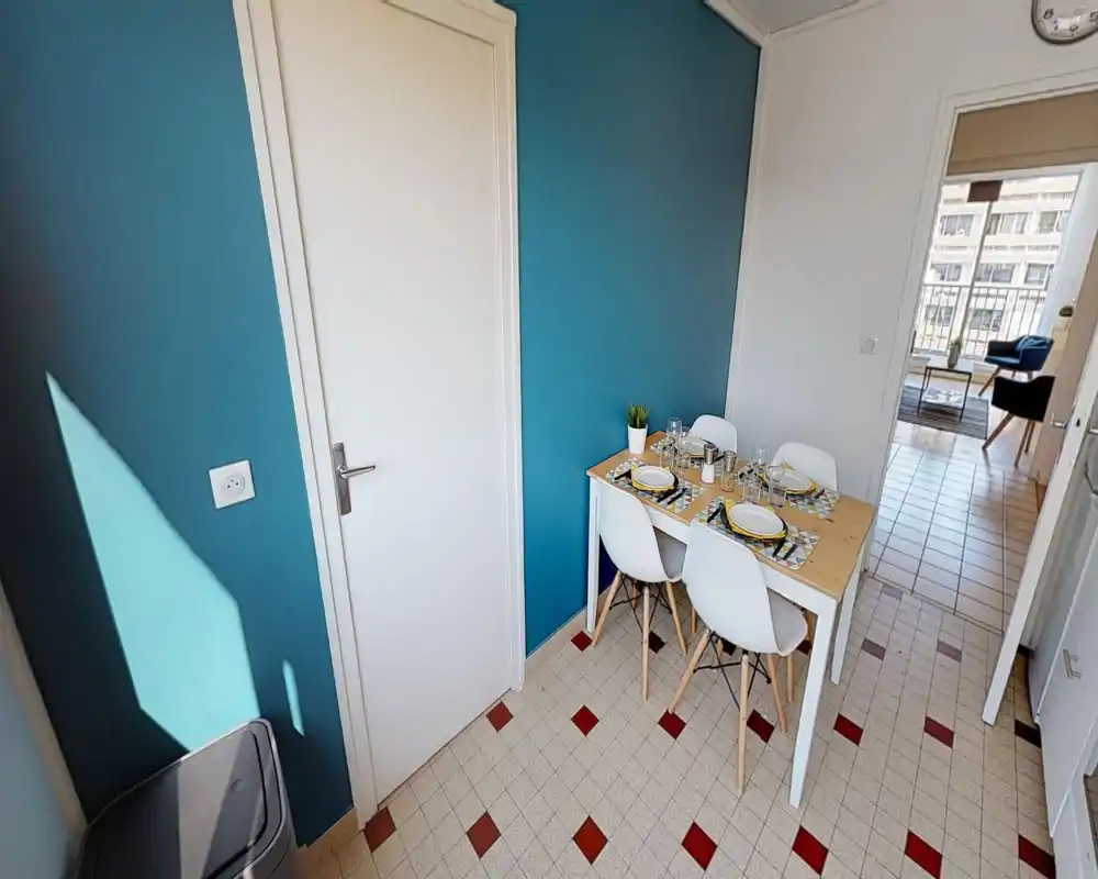 Petite salle à manger avec table en bois pour quatre personnes, chaises blanches modernes et sets de table colorés ; lumineuse et reliée au salon par une porte ouverte.