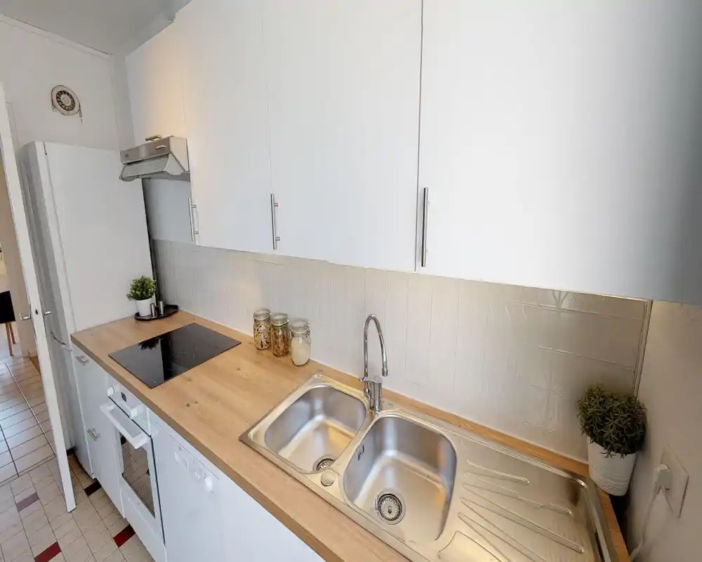 Cuisine moderne et compacte avec évier double en acier, plaque à induction et plan de travail en bois; meubles blancs et petits bocaux décoratifs apportent une ambiance lumineuse et soignée.
