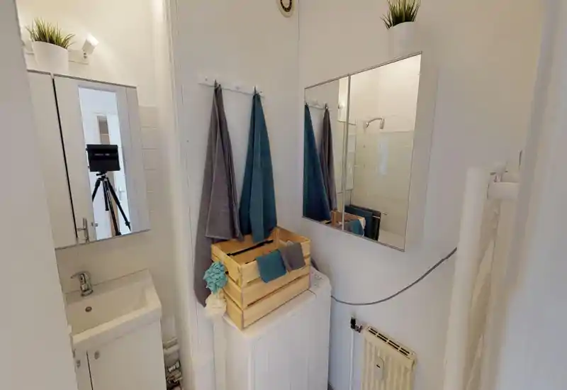 Bagno compatto e luminoso con piccolo lavabo, mobiletto con specchio, parete doccia e asciugamani appesi — ordinato e funzionale per gli ospiti.