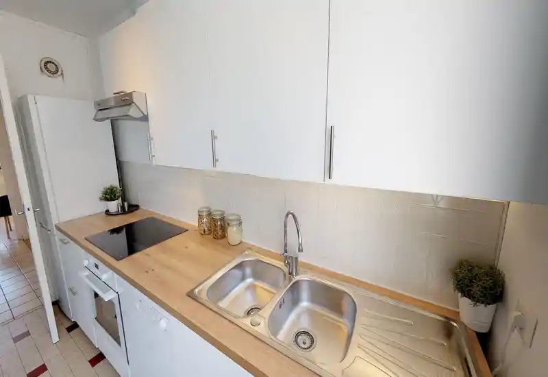 Cucina moderna compatta con doppio lavello in acciaio, piano cottura a induzione e piano in legno; mobili bianchi e piccoli barattoli decorativi con piante rendono lo spazio luminoso e ordinato.